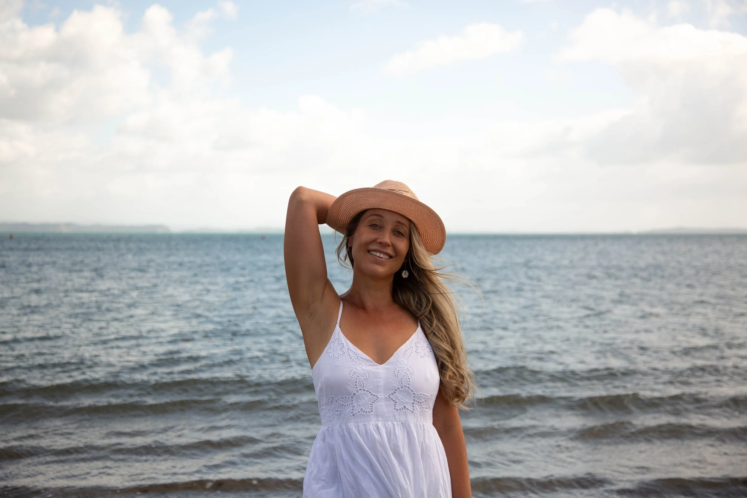 A woman in a white dress and a wide-brimmed hat stands on a beach near the water, smiling at the camera with her hand resting on her head, with a cloudy sky in the background.