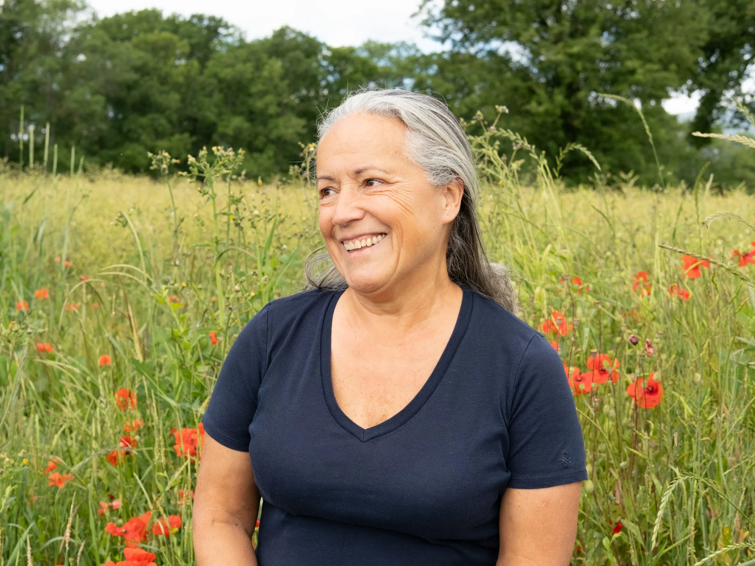 Une femme souriante portant un t-shirt bleu marine dans un champ de coquelicots et des arbres en arrière-plan