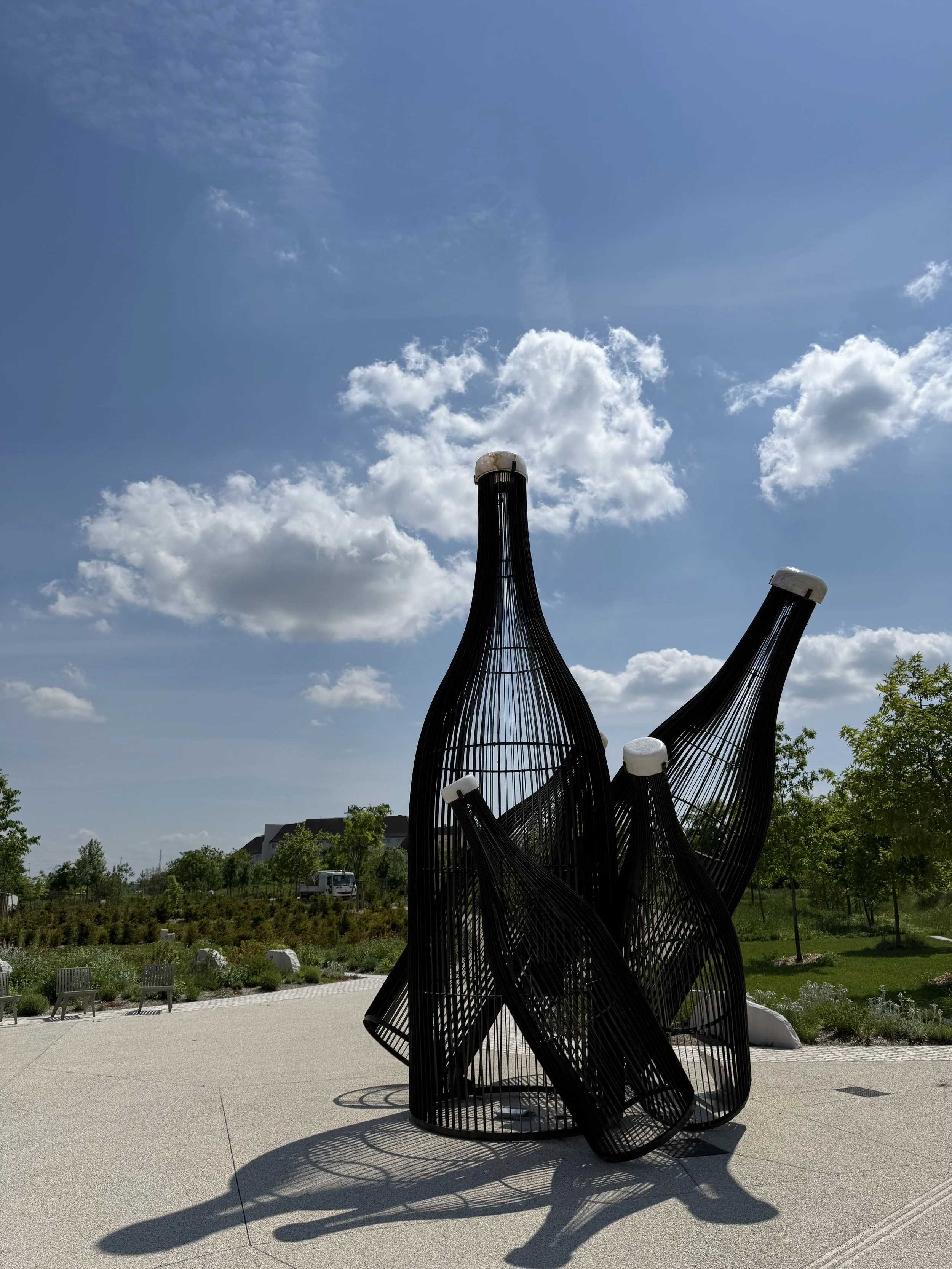 Kunstinstallation aus schwarzen, runden, undösierten Strukturen mit weißen Kopfbedeckungen, im Freien unter blauem Himmel mit Wolken, umgeben von Bäumen und Wiesen.