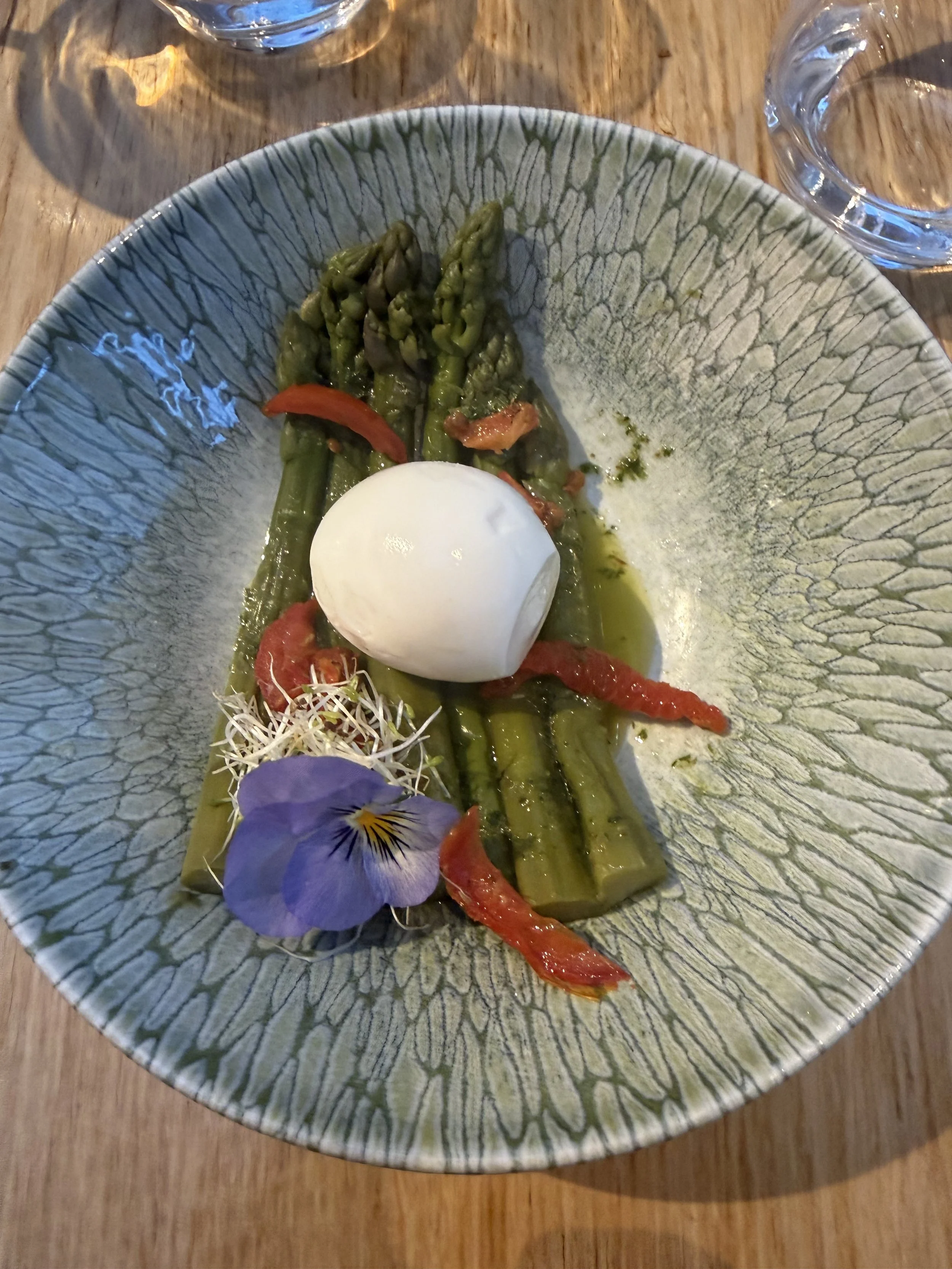 Grüner Spargel auf einem Teller mit einem pochierten Ei, roten Paprikastreifen, mikrozwei, Blüten und essbaren Blättern, serviert auf einem Holztablett.