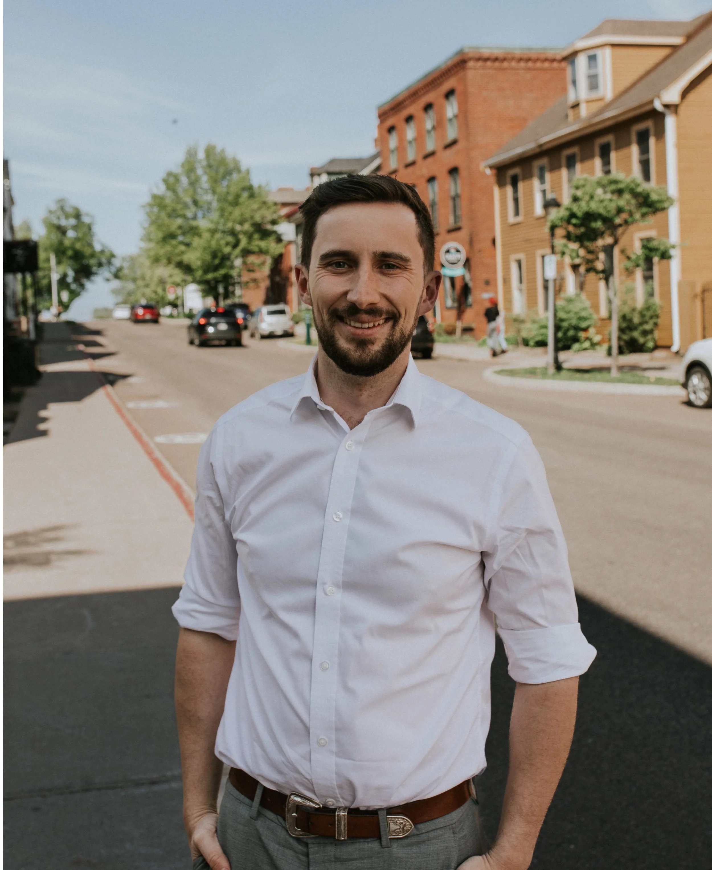A man with dark hair and a beard smiling wearing a white dress shirt with sleeve rolled up, gray trousers, and a brown belt, standing outdoors in a small town with brick and wooden buildings, parking spots, and trees in the background.
