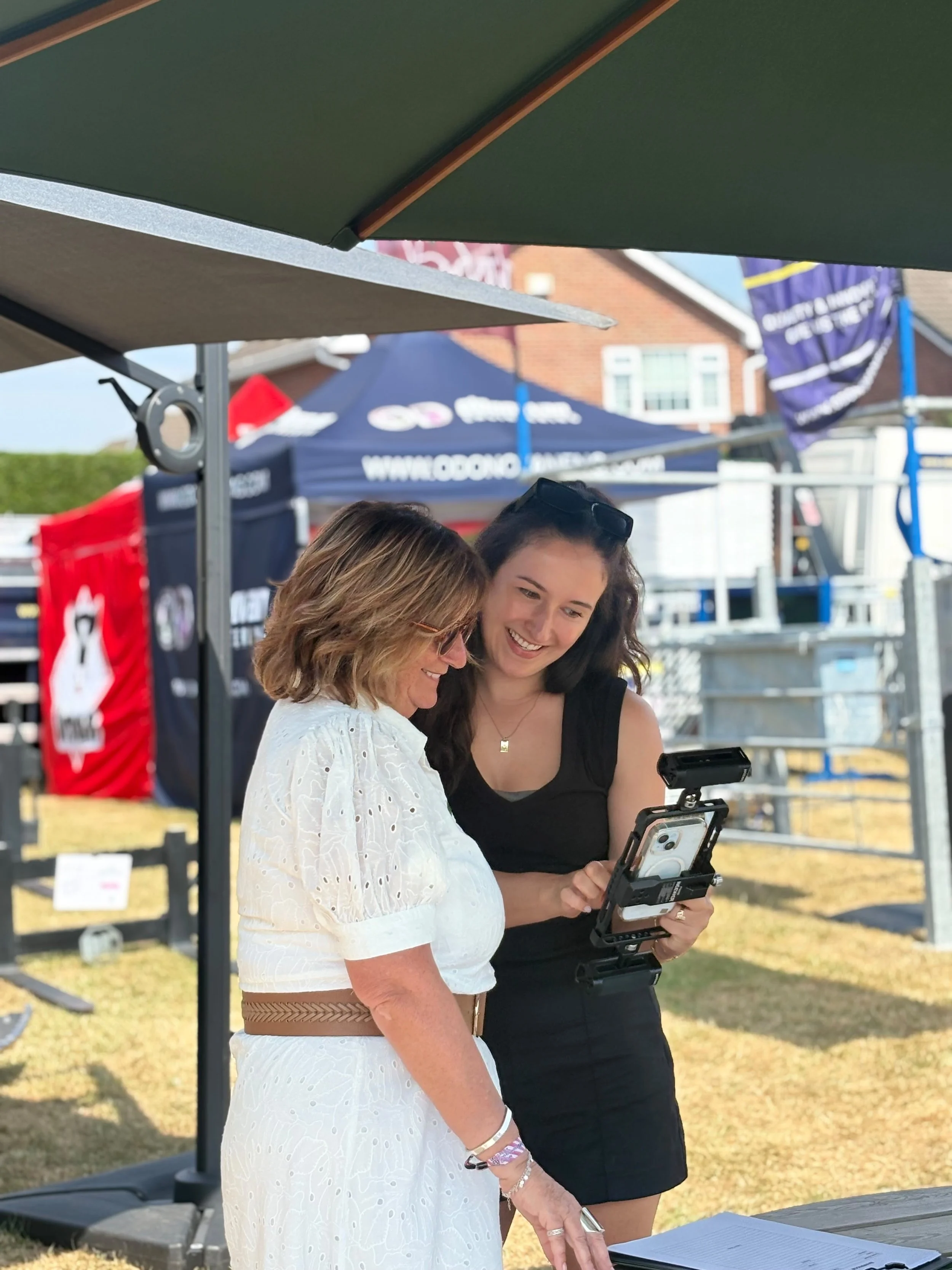 Two women engaging with a mobile device at an outdoor event, with tents and a building in the background.