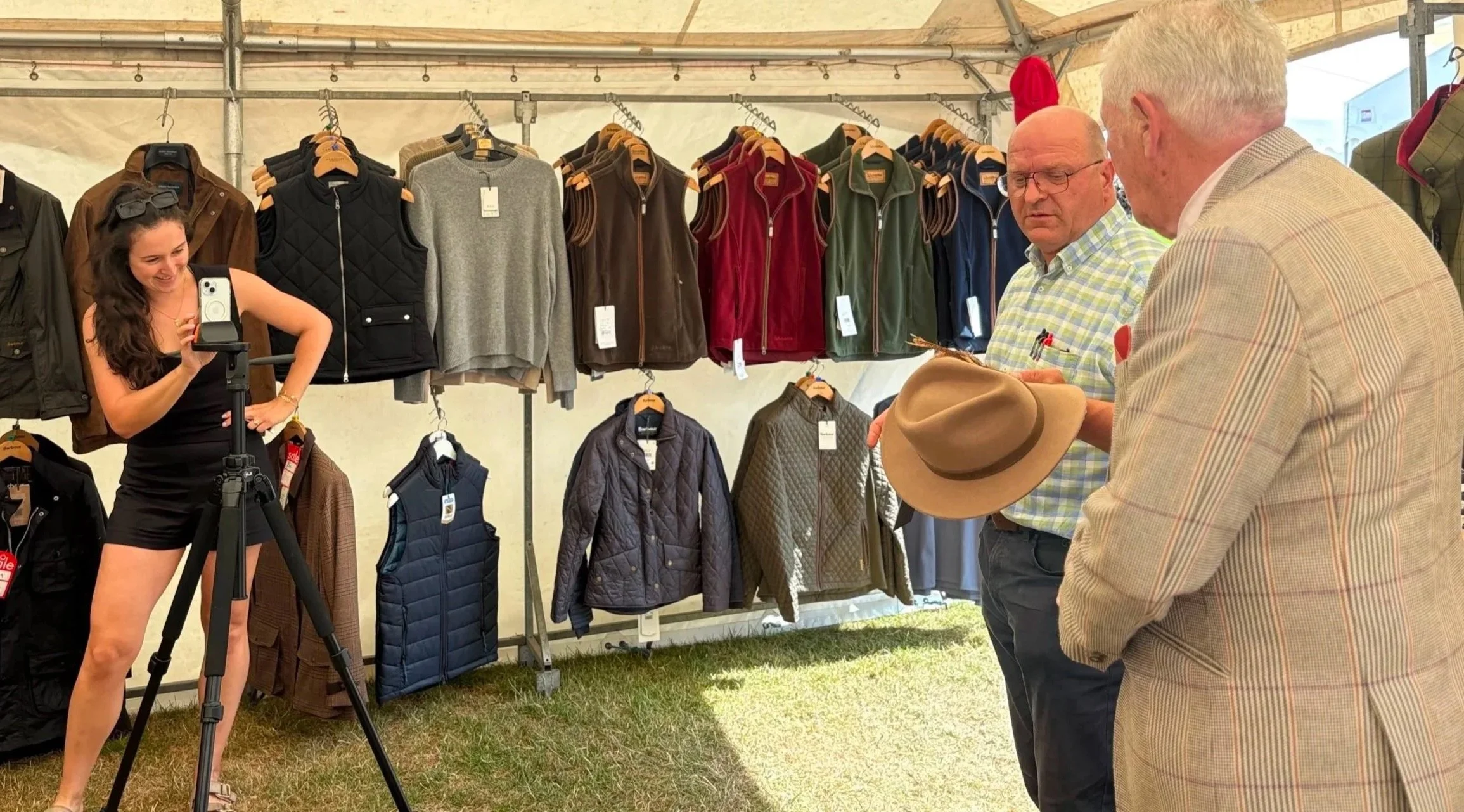 A woman filming a man and another man at a clothing stall at an outdoor event. The stall displays jackets, vests, and sweaters hanging on a rack under a tent. The woman has long curly hair and is wearing a black sleeveless top and black shorts. One man has white hair, glasses, and is wearing a plaid shirt, holding a hat. The other man, with blonde hair, is wearing a beige plaid blazer.