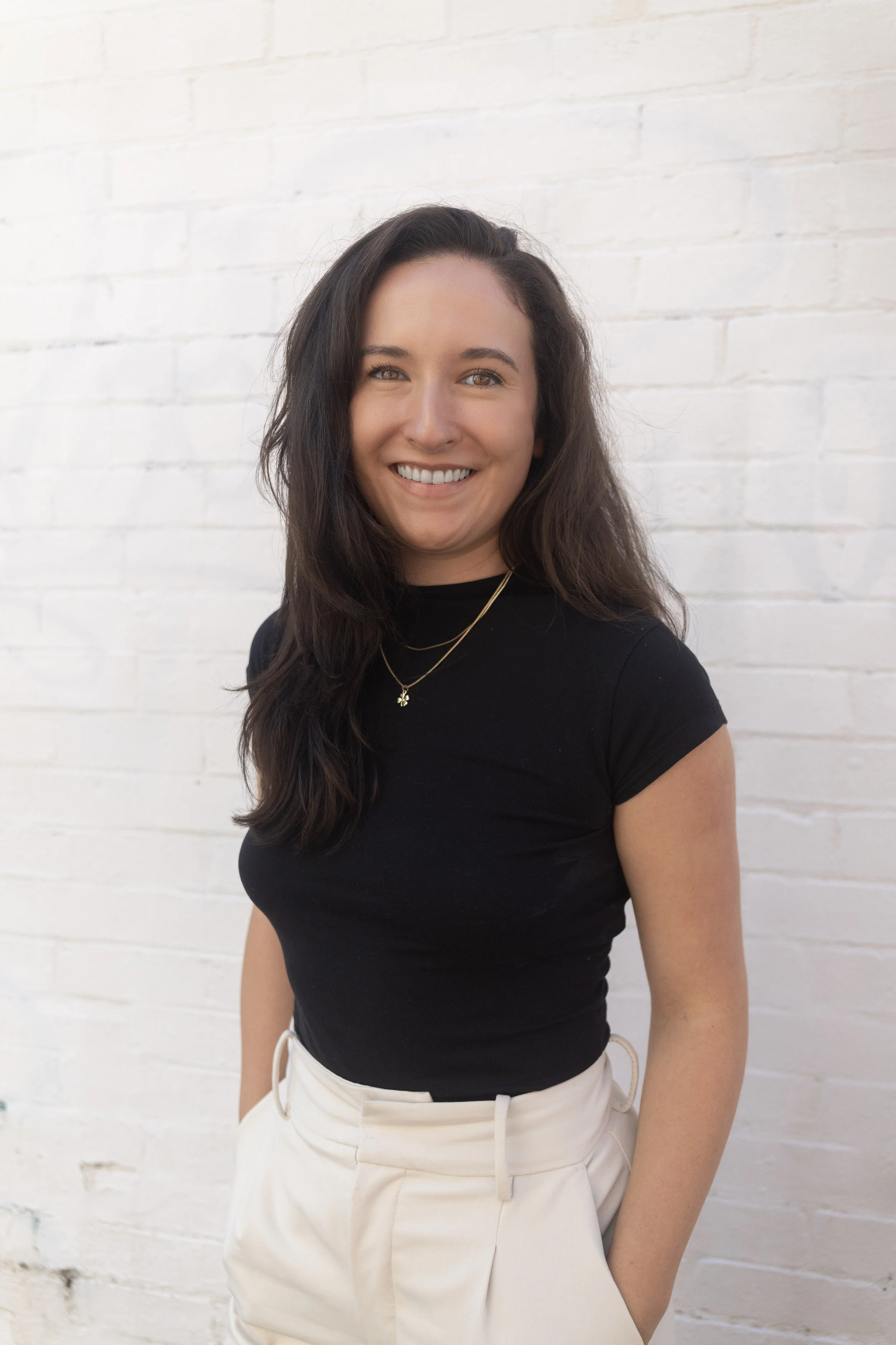 A woman with long, wavy dark hair smiling, wearing a black short-sleeved top, jewelry, and high-waisted cream-colored pants, standing against a white brick wall.