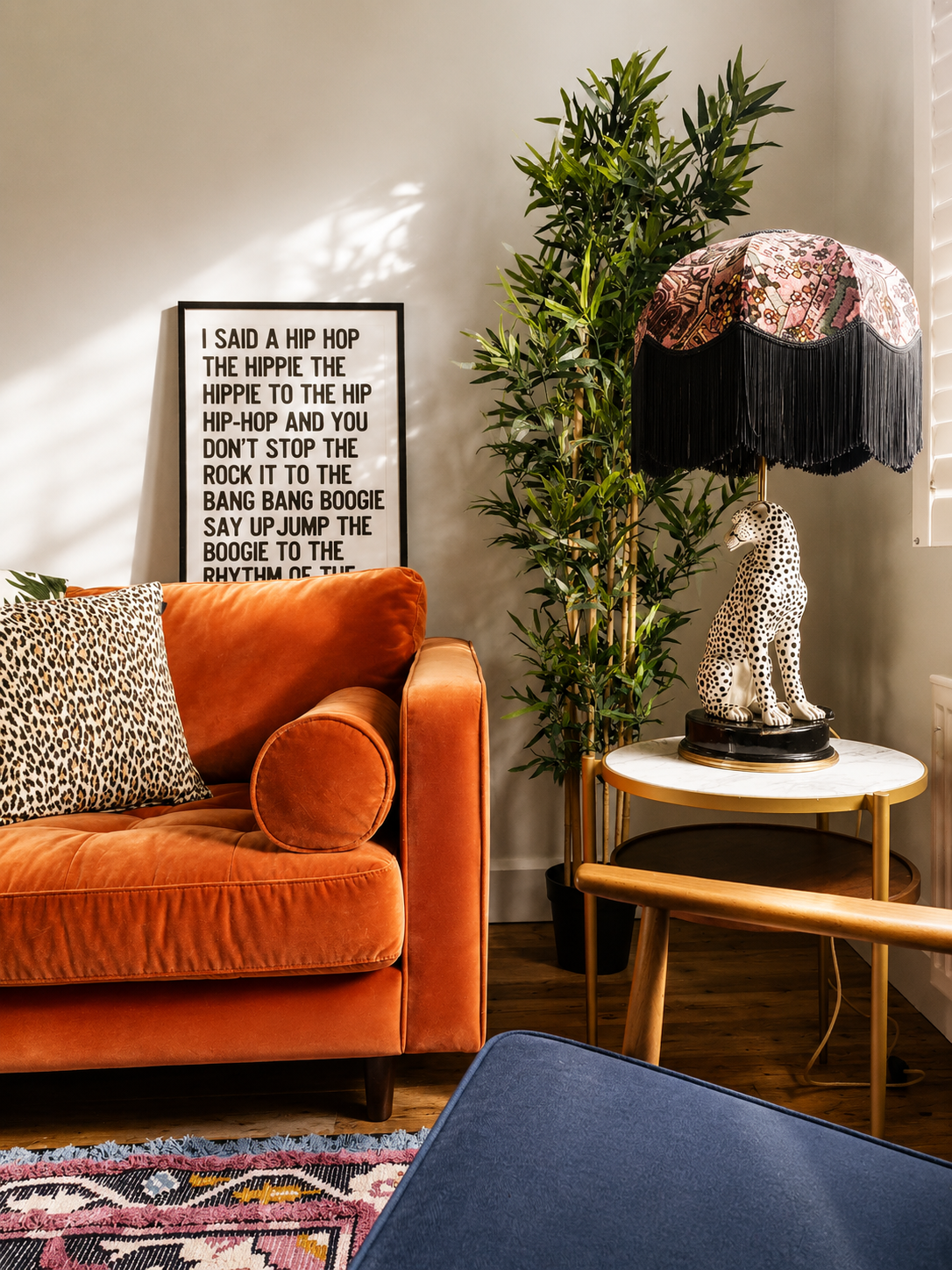 Living room corner with orange velvet sofa, leopard print pillow, framed text art, tall indoor plant, patterned lampshade on leopard figurine table, and blue fabric chair.