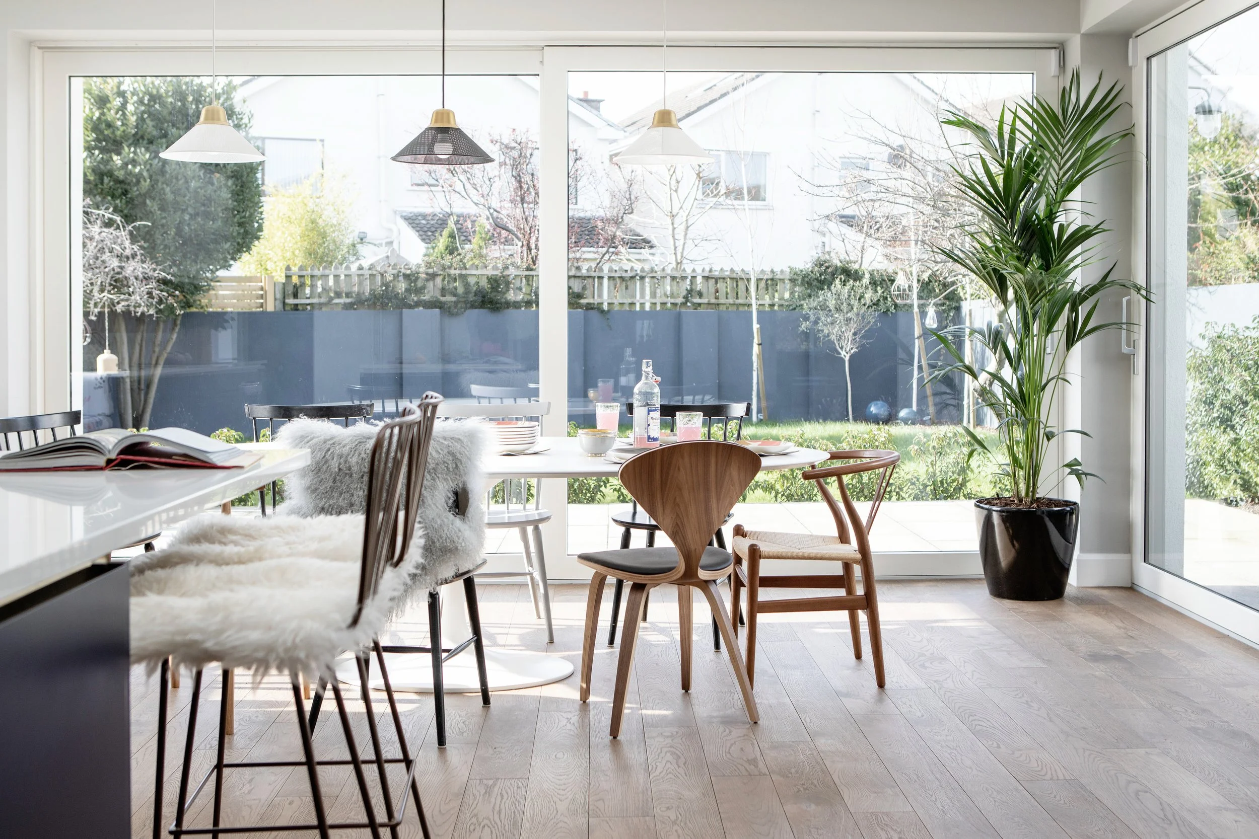 A bright indoor dining area with large glass sliding doors opening to a backyard garden. There is a round marble table with diverse chairs, a tall potted plant, and hanging pendant lights.