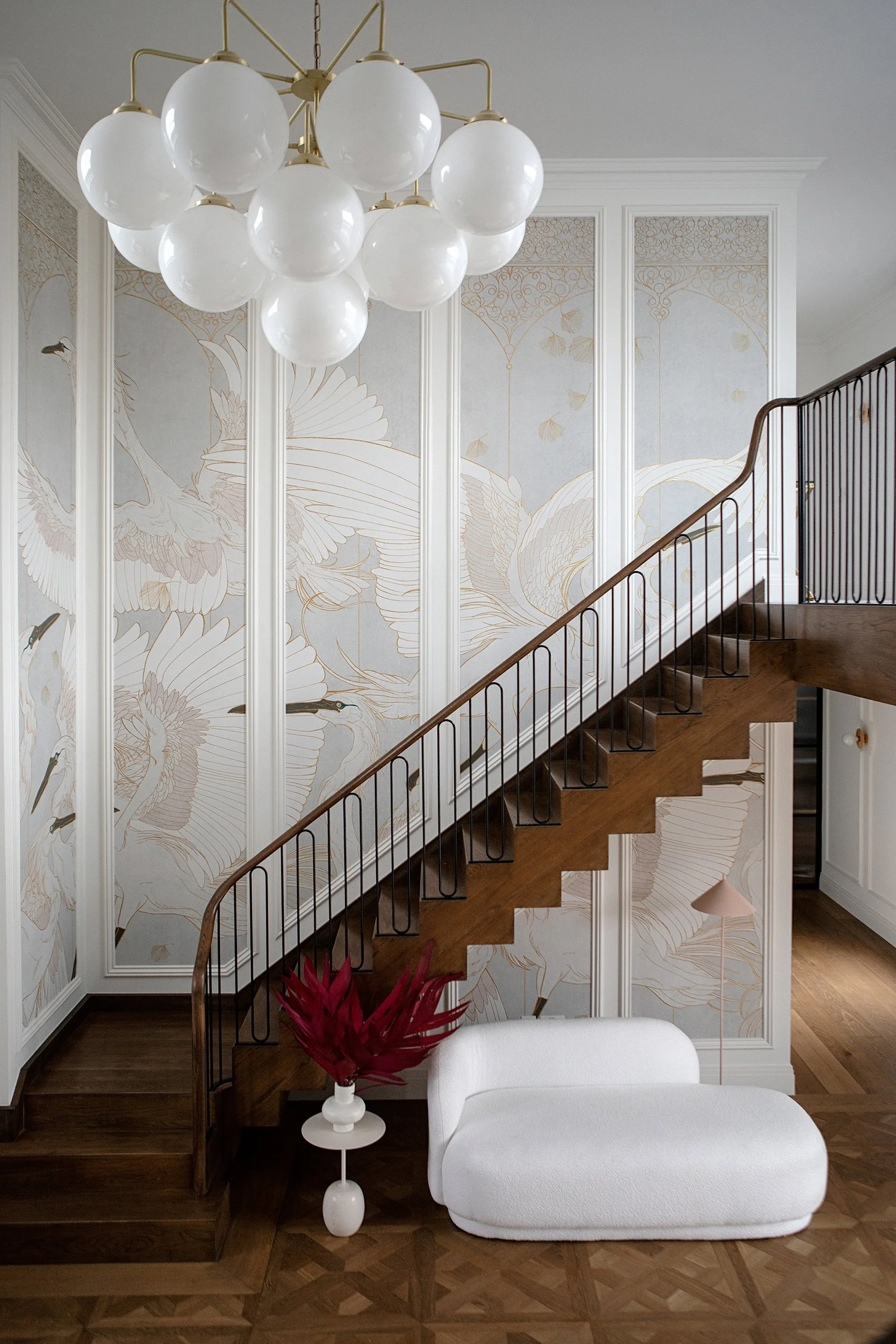 Interior view of a staircase with a modern chandelier in a huge double height entrance, white wall with panels and wallpaper, a white boucle chaise longue, and a red plant in a white vase.