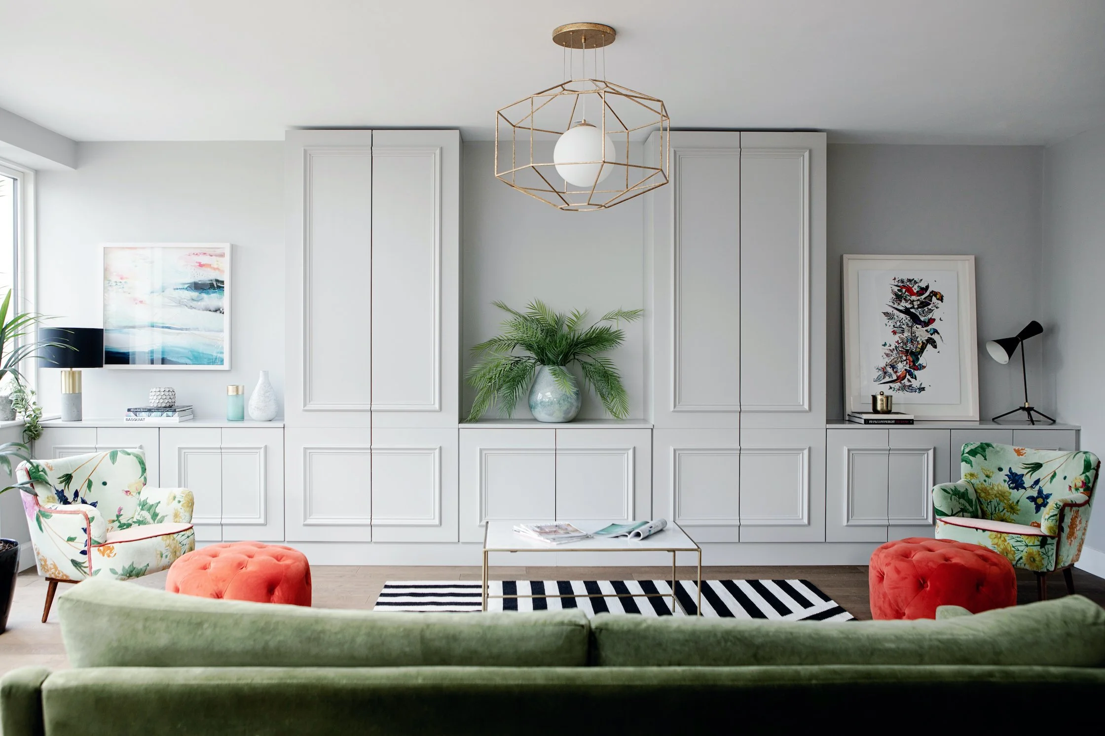 Modern living room with white built-in cabinets, colorful floral armchairs, a green velvet sofa, a black and white striped rug, a marble coffee table, artwork on the walls, potted plants, decorative lamps, and a geometric gold pendant light.