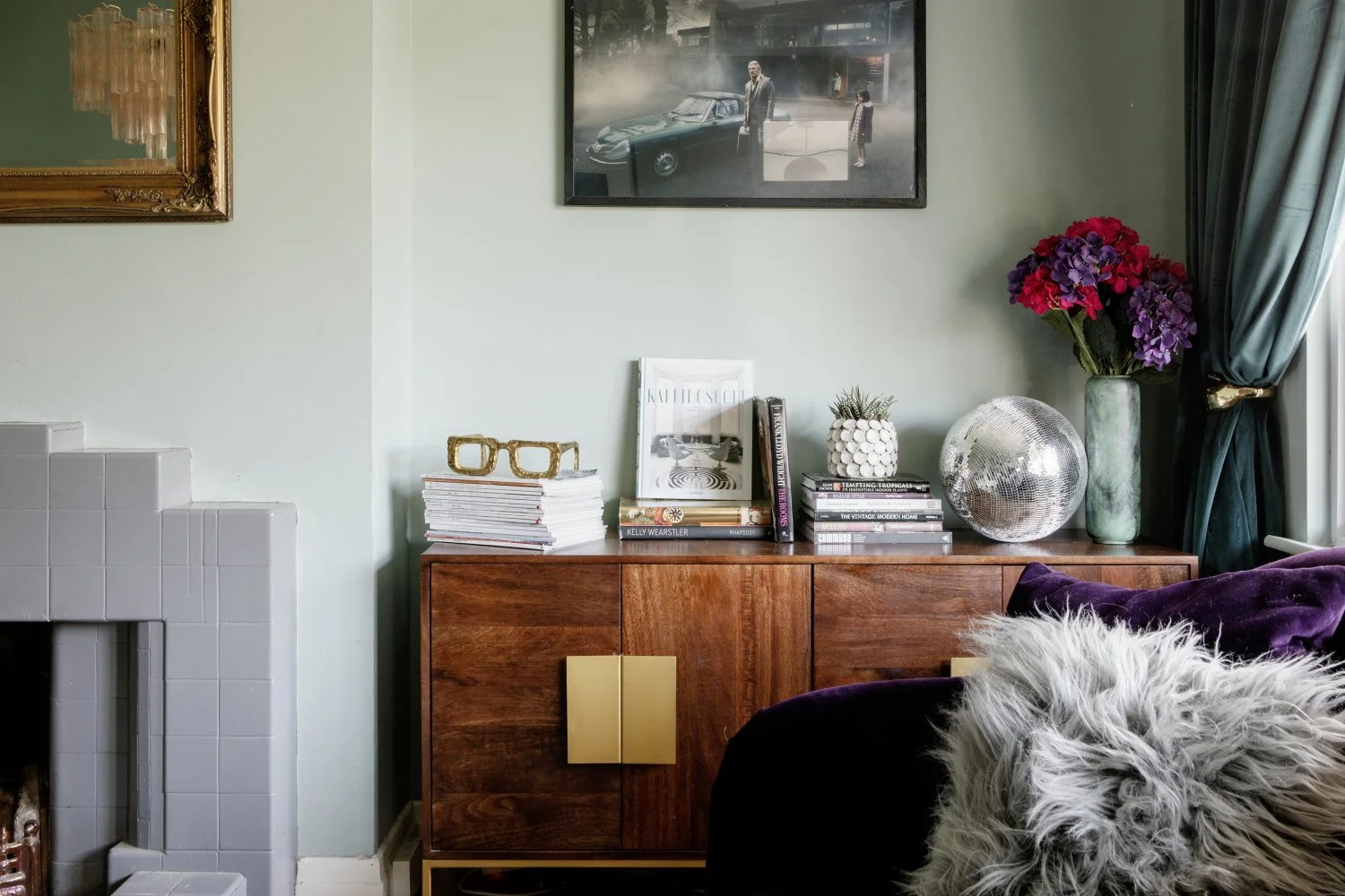 A living room corner with a wooden sideboard decorated with magazines, books, a white decorative pineapple, a disco ball, and vases with flowers. Two framed artworks hang on the pastel green wall, with a green curtain and purple velvet cushion visible on the right.