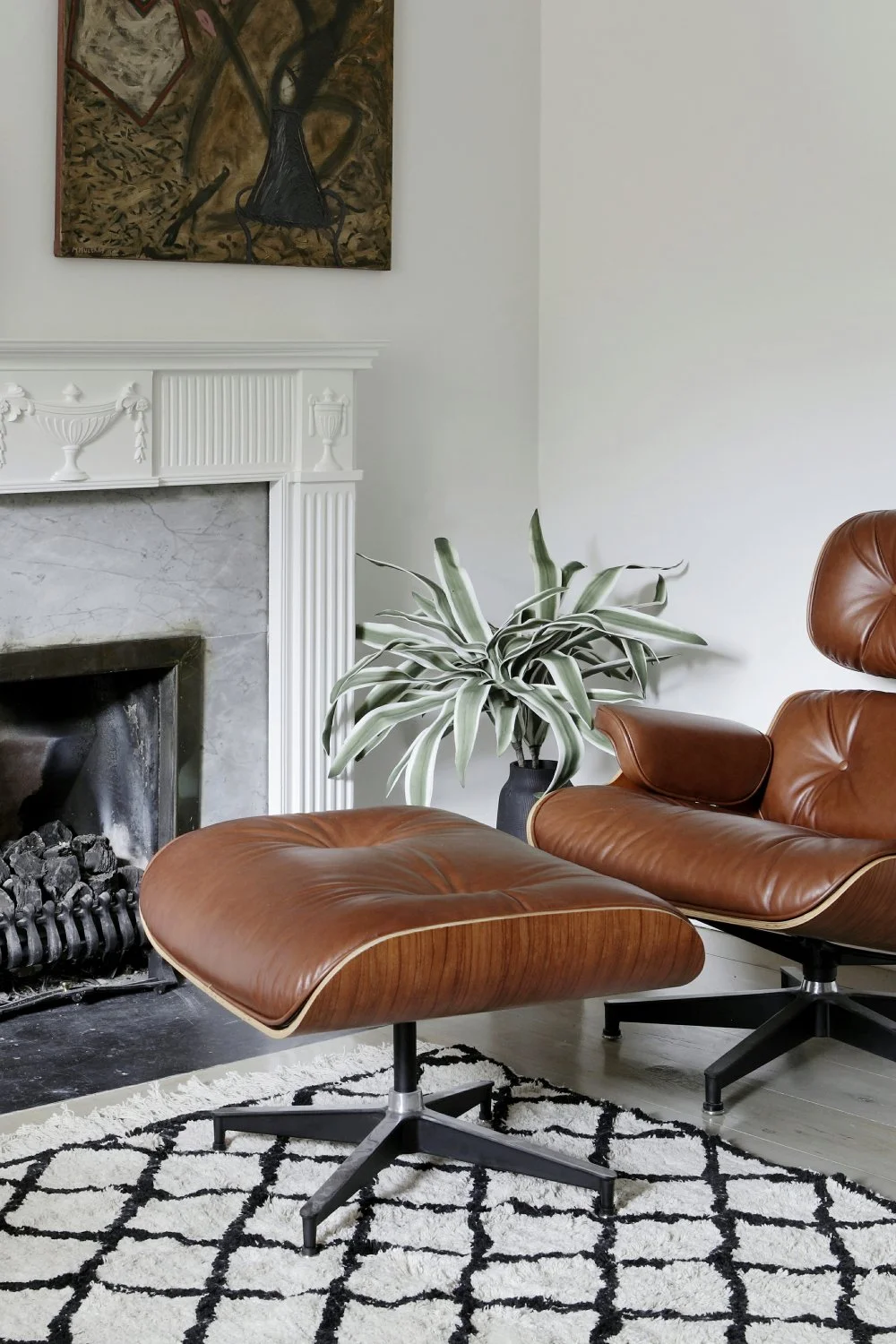 Living room with a leather lounge chair and ottoman, a potted plant, fireplace, a black and white rug, and artwork above the fireplace.