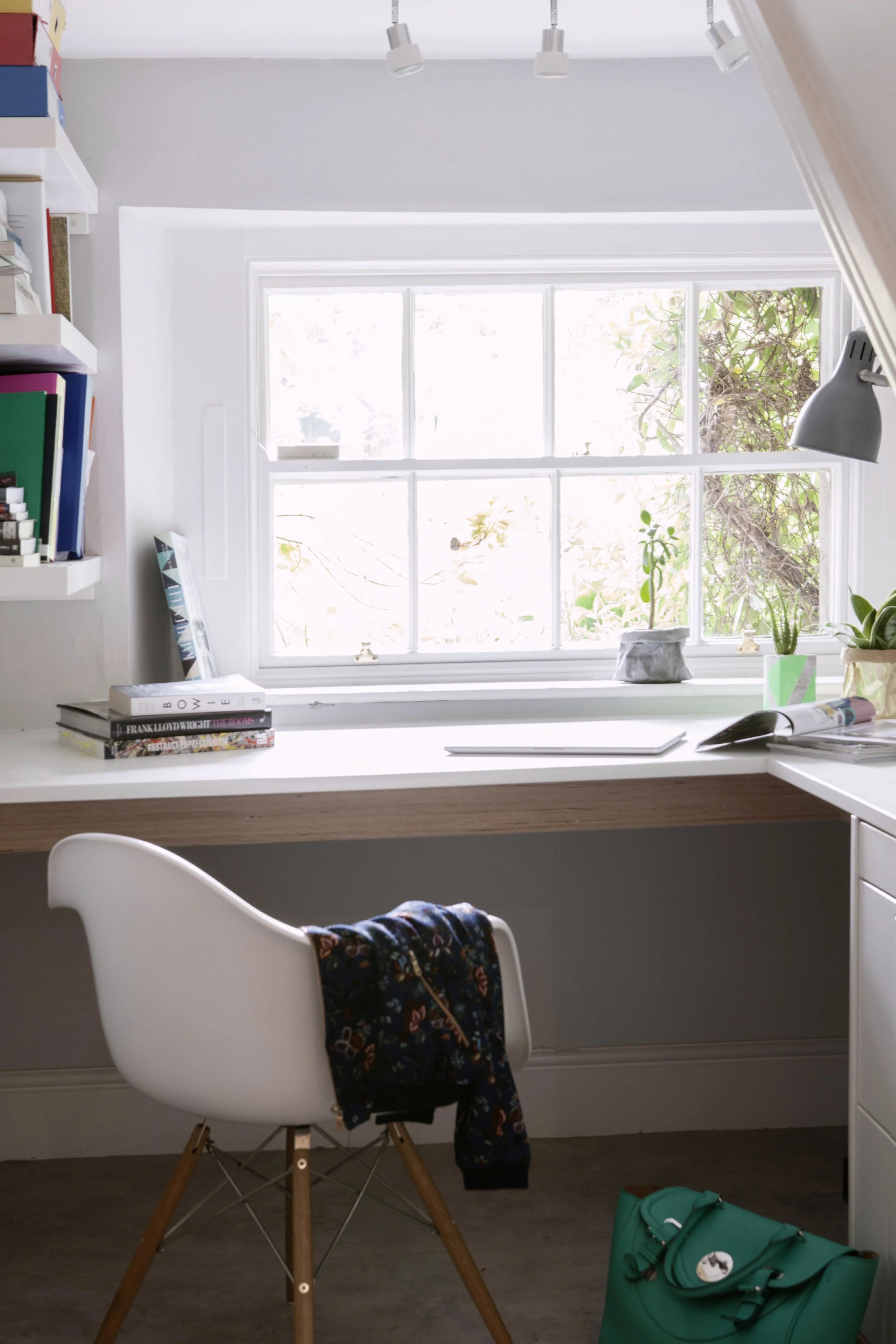 A home office workspace with a white desk, a white chair with wooden legs, a window with white frames, books on shelves, a potted plant on the desk, and a teal bag on the floor.