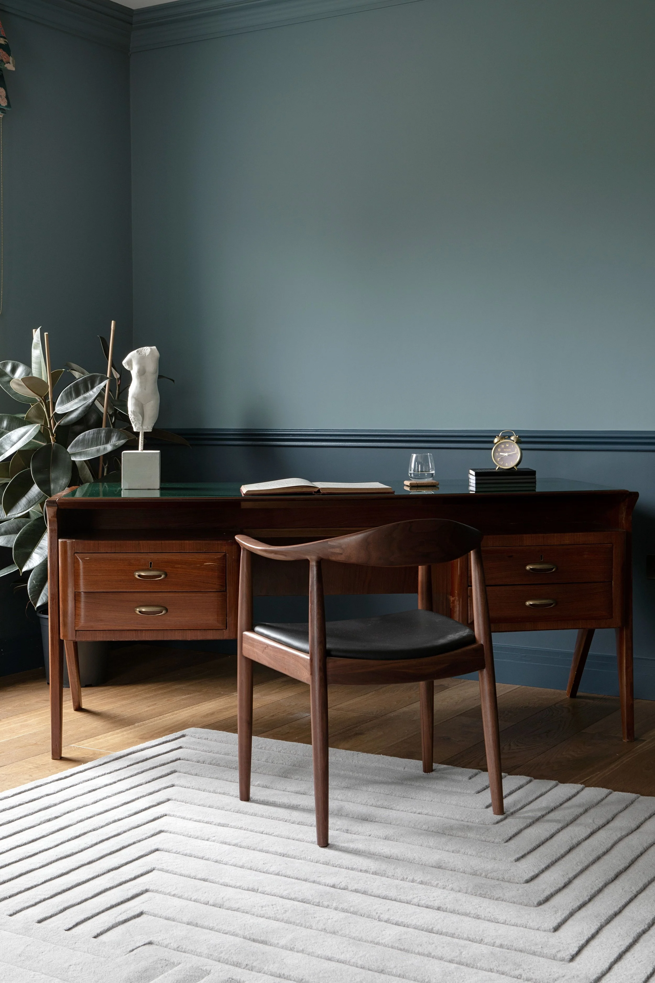 A wooden desk with a chair in a room with teal walls. The desk has a small clock, a glass of water, an open book, and a decorative sculpture of a female torso. A potted plant with large, glossy leaves is on the left side of the desk. There is a light-colored striped rug on the wooden floor.