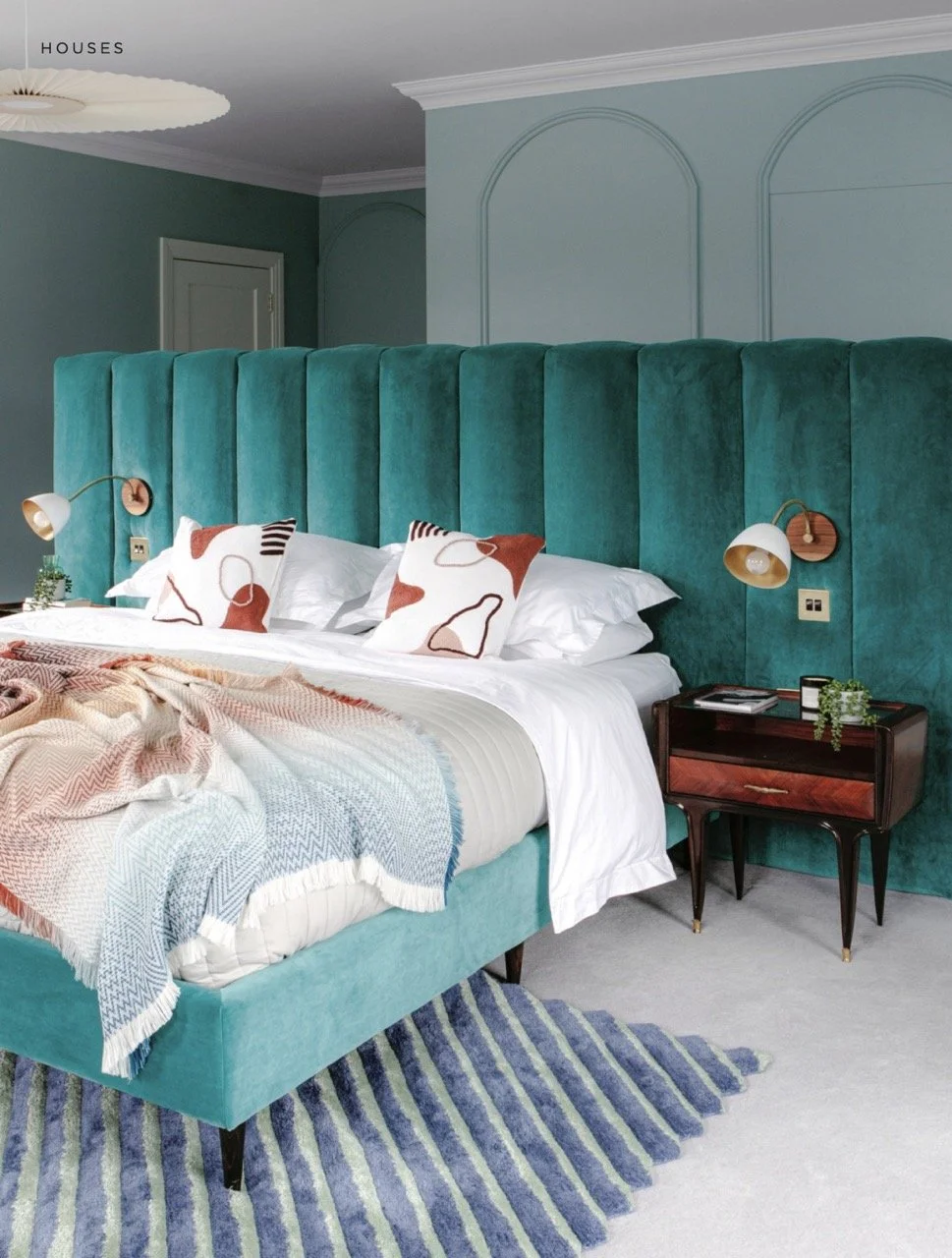 Modern bedroom with a teal velvet headboard, white bedding, and patterned pillows. A striped rug is partially visible on the light carpeted floor. A dark wood bedside table holds a small plant, books, and a candle, with wall-mounted reading lamps on either side of the headboard.