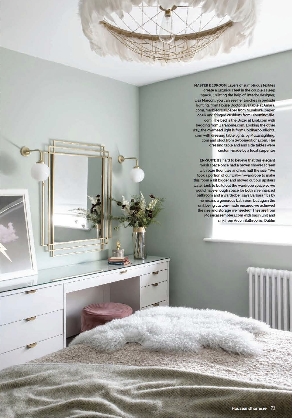 A bedroom with a pastel green wall, a white dresser with gold handles, a mirror with a gold geometric frame, two wall-mounted spherical light fixtures, and a floral arrangement in a glass vase. The bed has a beige blanket and a white fluffy throw, with a pink velvet stool in front of the dresser. The ceiling features a decorative paper lantern with a gold interior.