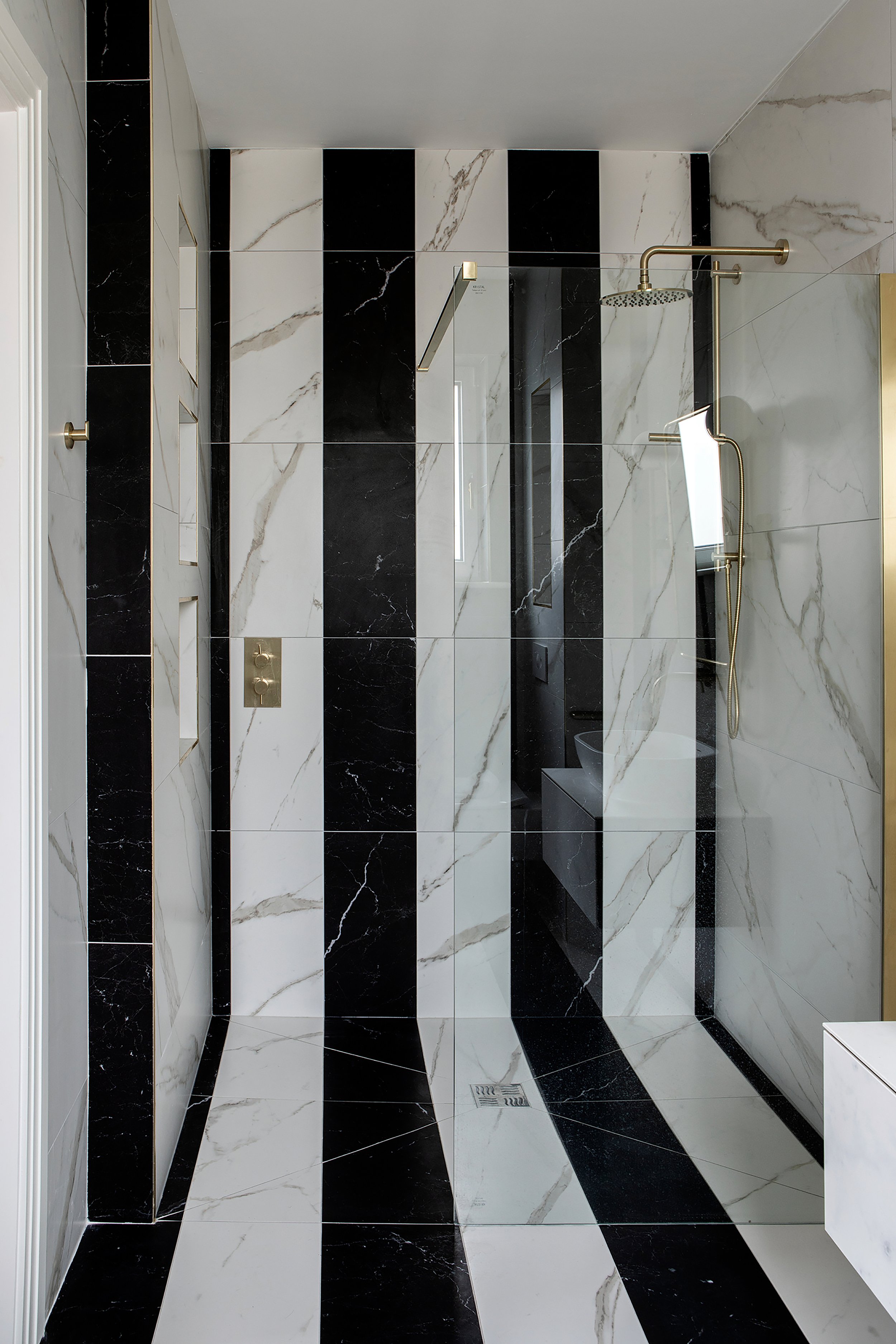 A modern shower area with black and white marble tiles arranged in vertical stripes on the walls and horizontal stripes on the floor. The shower has a gold-rimmed rainfall showerhead, a handheld shower wand, and a control panel with gold accents. There's a small window and a drain on the floor.