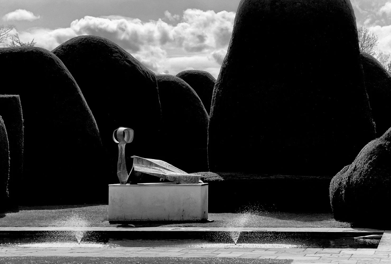 Sculpture of a reclining figure on a pedestal in front of large, rounded bushes and a cloudy sky.