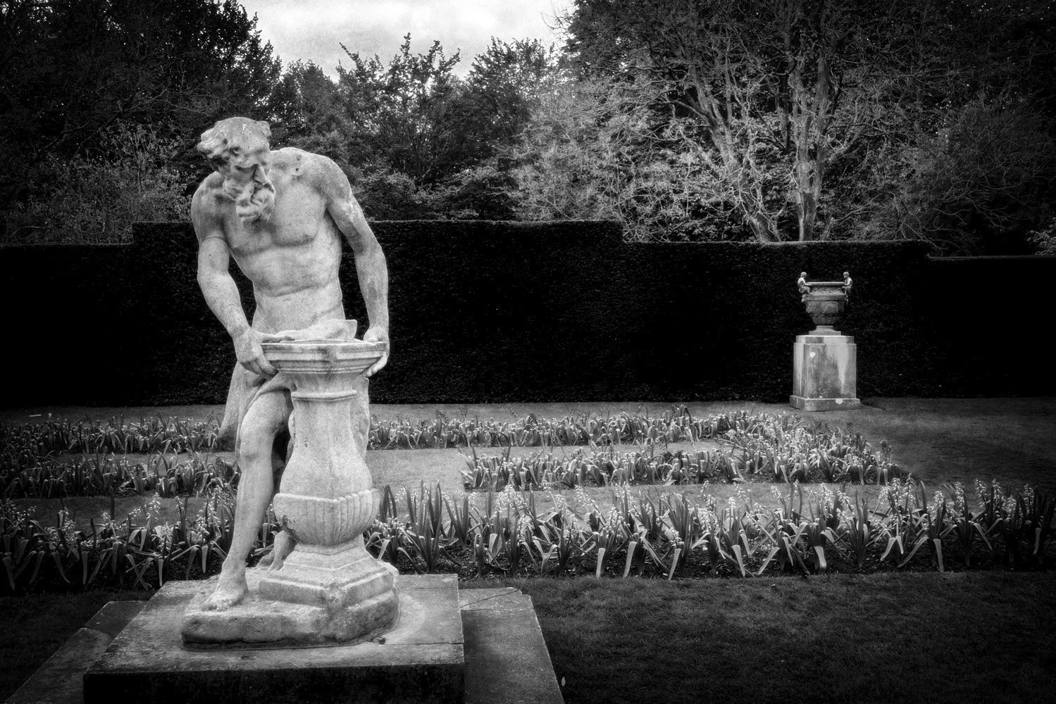 Black and white photo of classical sculpture of Father Time in a garden with trees and flowers.