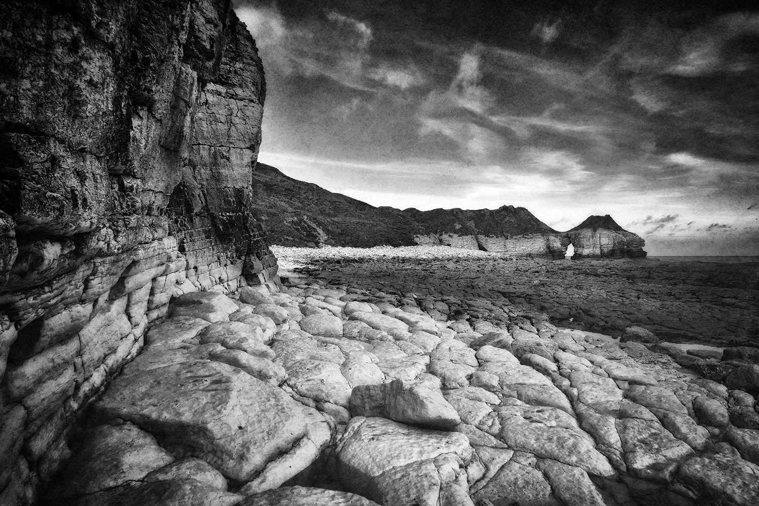 Black and white photograph of a rocky seashore with steep cliffs on the left, textured rocks on the ground, and distant cliffs with a natural arch formation near the horizon, under a dramatic cloudy sky.