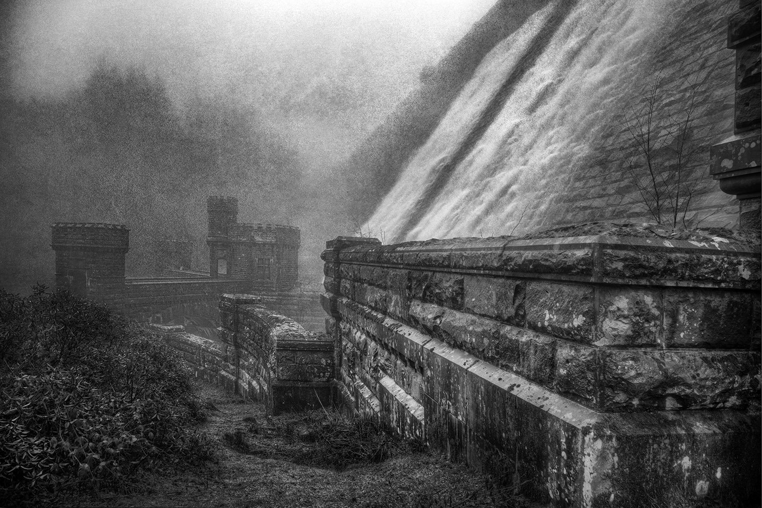 Black and white photo of an old stone bridge with castle-like towers in the background, some small trees and shrubbery next to the bridge, cloudy sky.