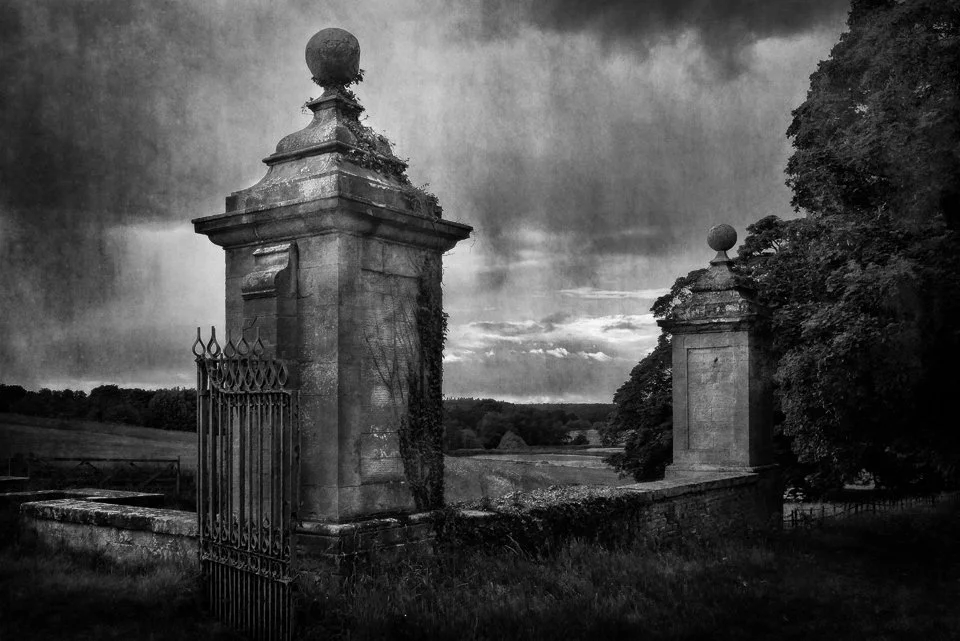 Black and white photograph of old stone pillars with decorative iron gate, surrounded by trees and fields, under a cloudy sky.