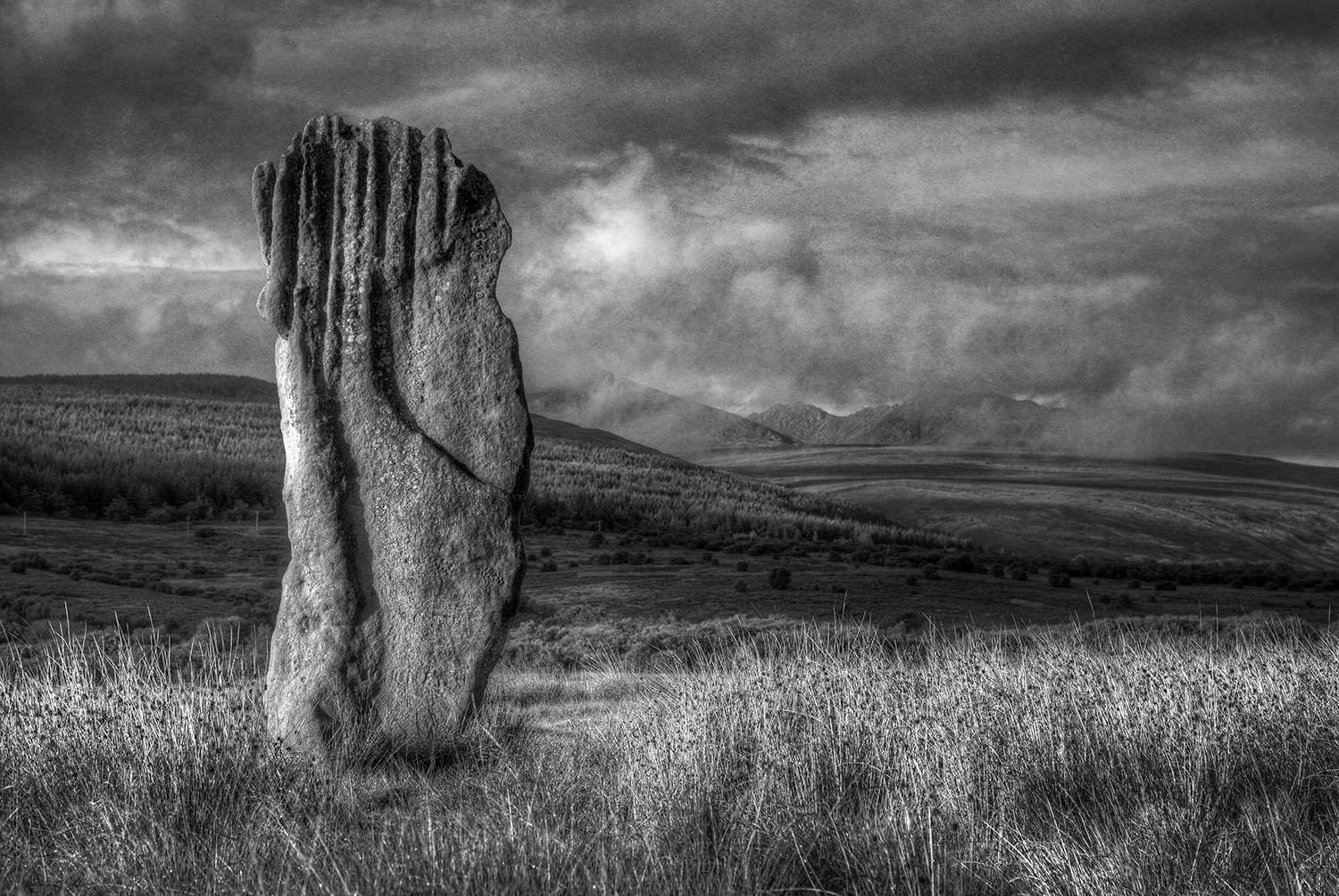 A large standing stone in a grassy field with mountainous terrain and clouds in the background, in black and white.