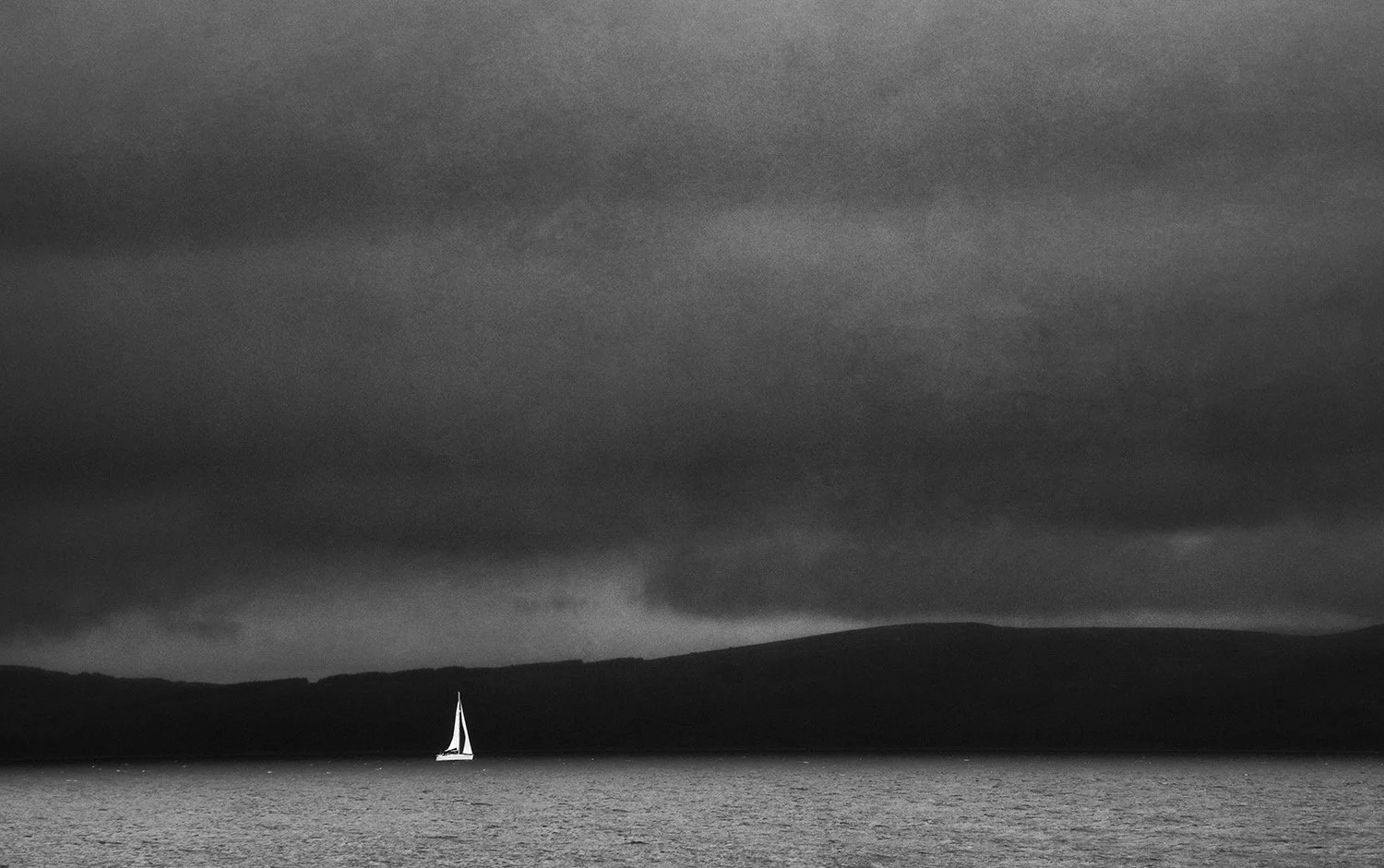 A small sailboat with a white sail on dark water under a stormy, cloud-covered sky