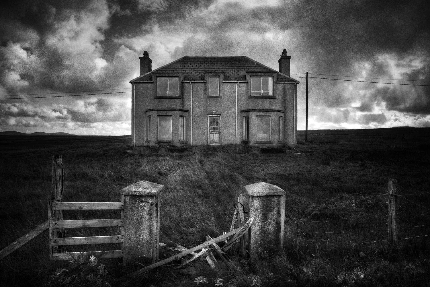 A black and white photo of an abandoned house on a hill under a cloudy sky, with a broken wooden gate in the foreground.