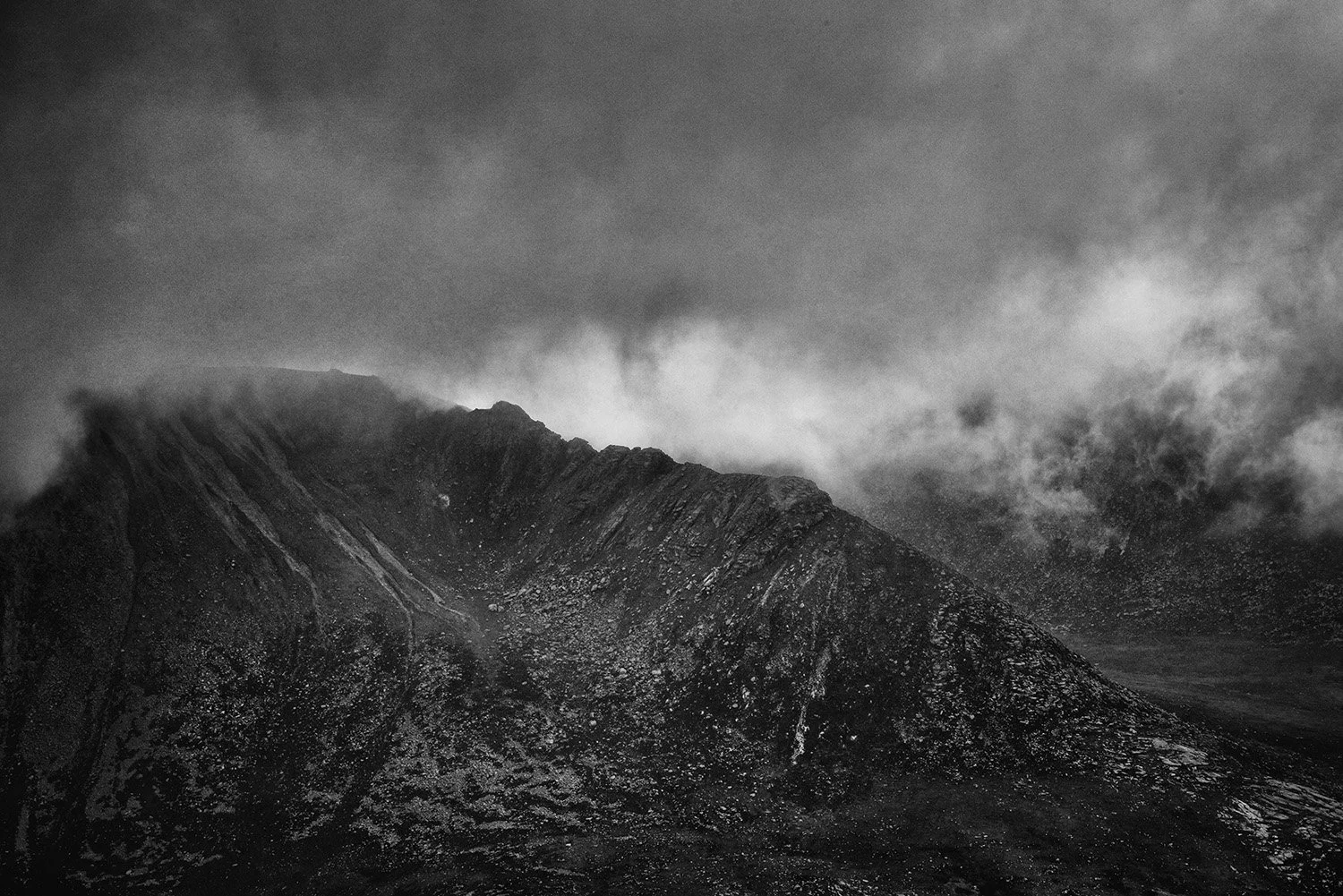 Black and white photo of a mountain peak partly covered by clouds, with rocky slopes and rugged terrain.