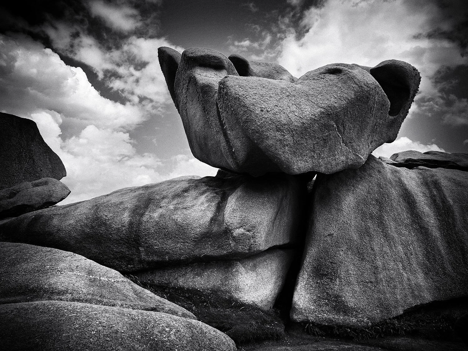 Black and white photo of large granite rocks, looking like two nuzzling dogs, stacked on top of each other against a cloudy sky.