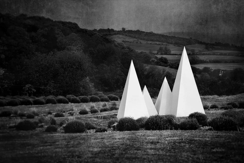 Black and white photo of a landscape with rolling hills and trees in the background. In the foreground, there are four large white pyramid sculptures standing out against a stormy sky.