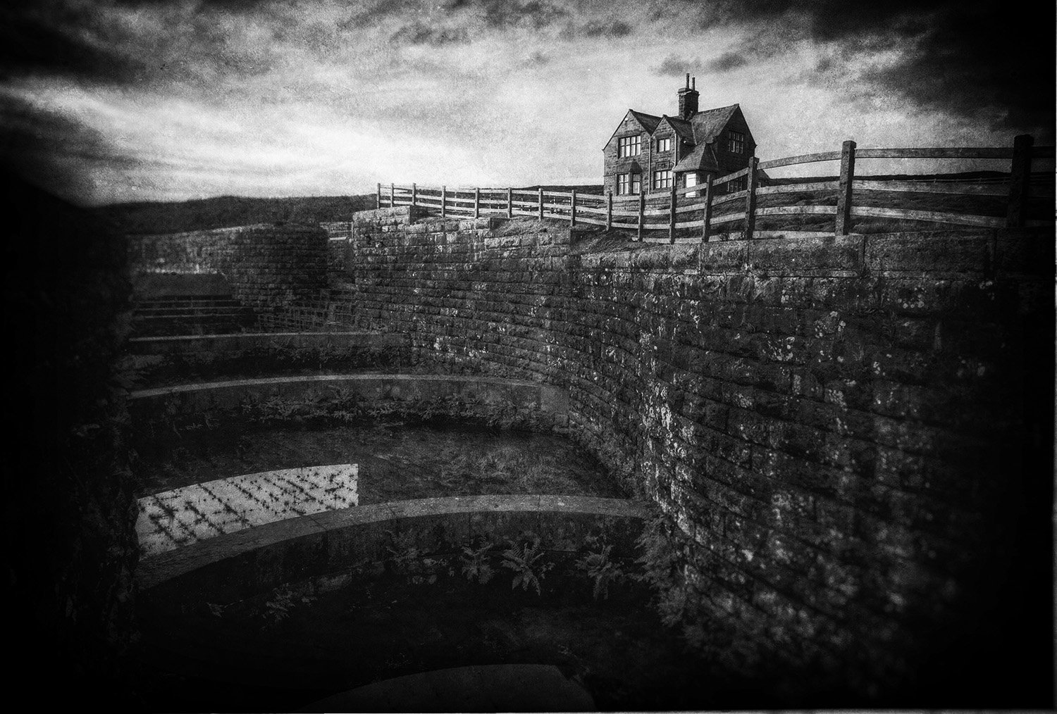 Black and white photograph of an atmospheric victorian reservoir building, surrounded by a stone wall, and a house on a hill in the background under a cloudy sky.