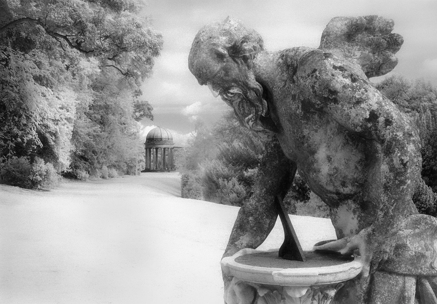 Black and white photo of a weathered stone sculpture of Father Time, with a beard, leaning forward over a sundialt, surrounded by trees and a classical pavilion in the distance.