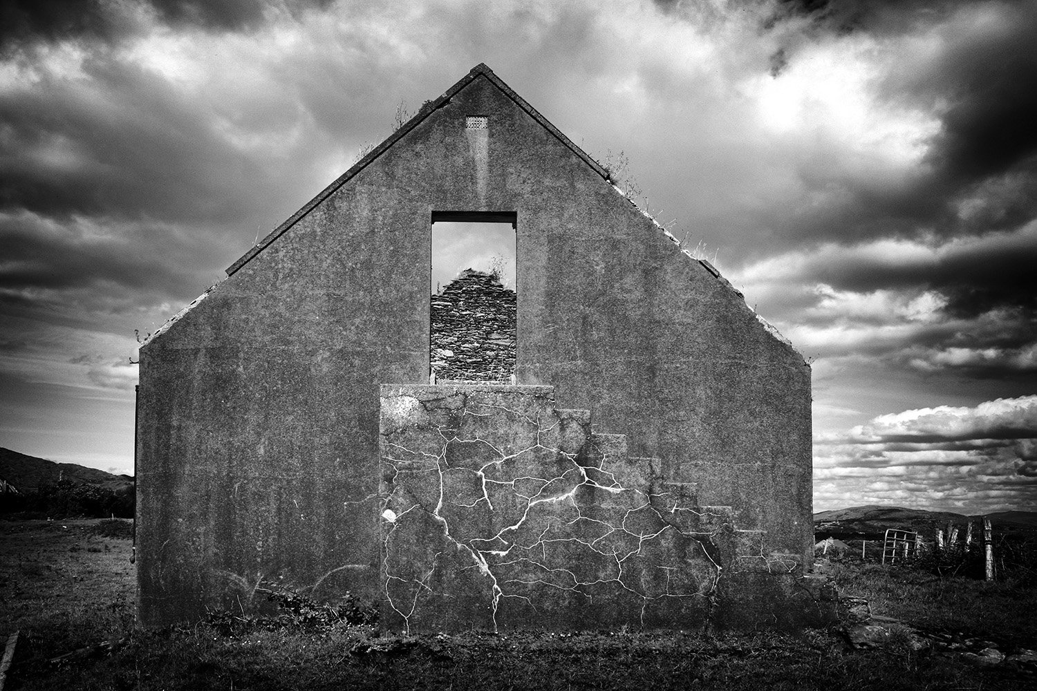 Black and white photo of an abandoned house with cracked walls, missing windows, and a partially collapsed roof, set against a cloudy sky.