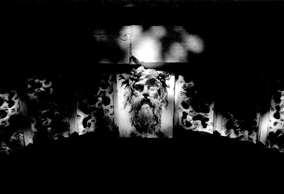 Black and white photograph of the central figure of a classical sculpture of a bearded man with a crown of leaves, framed by a decorative border with skull motifs, with a bright light source above illuminating the scene.