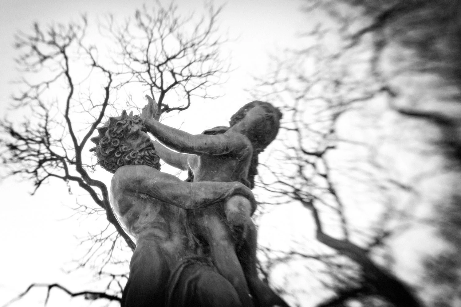 Black and white photograph of a statue depicting a muscular man with a beard and curly hair, wrestling with a mythological creature, set against the backdrop of leafless trees.