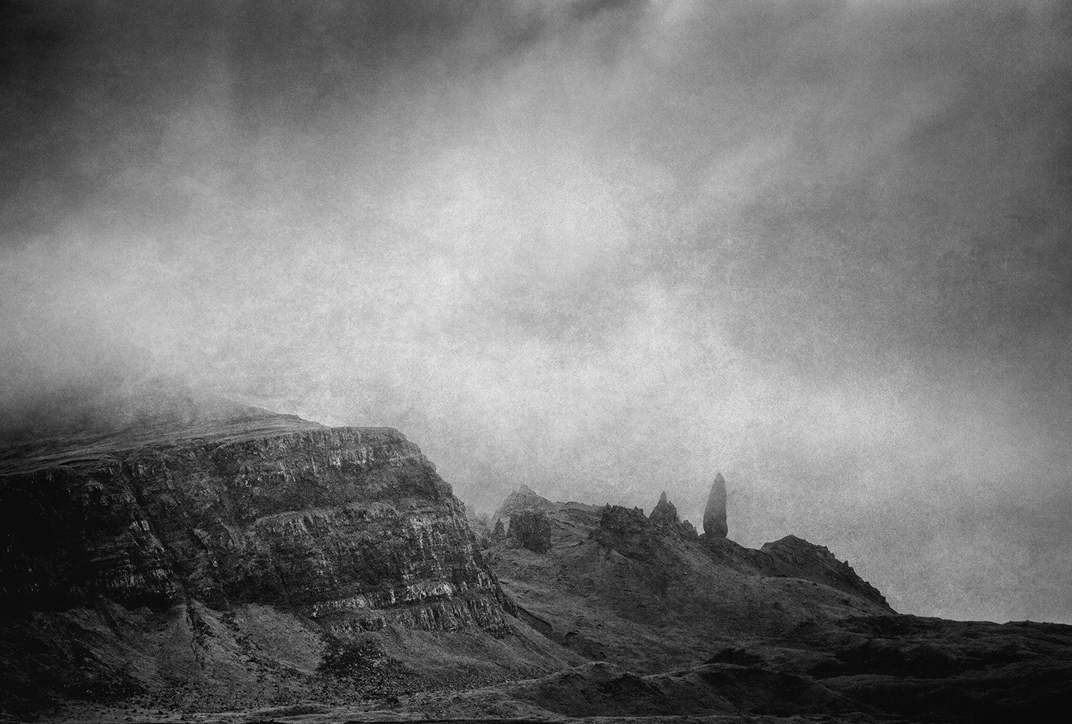 A black and white photograph of rugged mountain cliffs with sharp rock formations and a cloudy, misty sky overhead.