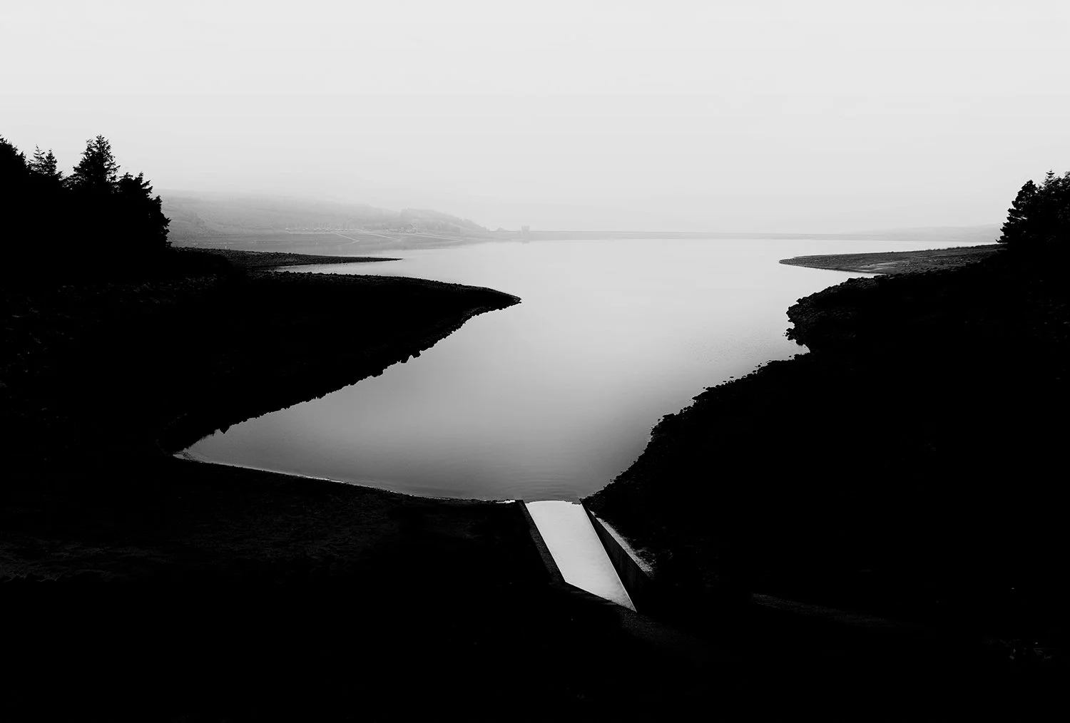 Black and white photo of a calm  reservoir with a small water spillway at the bottom center, surrounded by trees and hills in the distance.