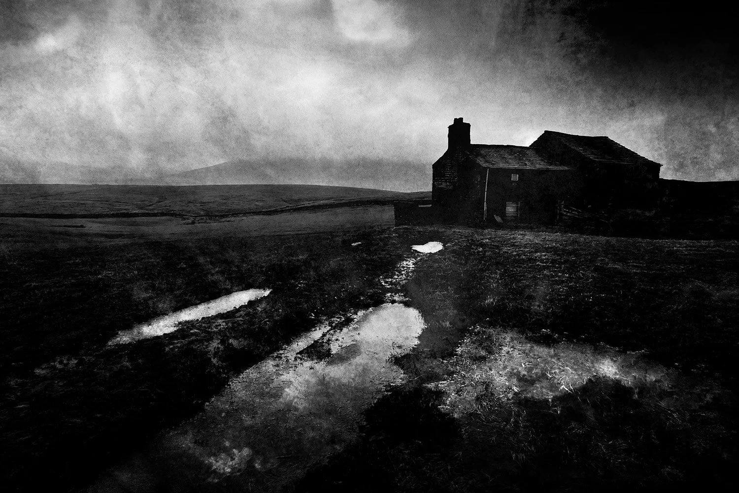 Black and white image of a solitary house with a chimney on a barren landscape, dark cloudy sky overhead, and puddles of water on the muddy ground.