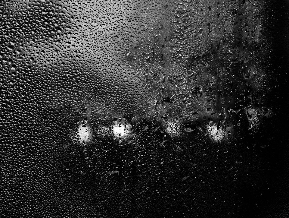 Close-up of a wet glass window with raindrops, with blurry city lights visible through the glass.