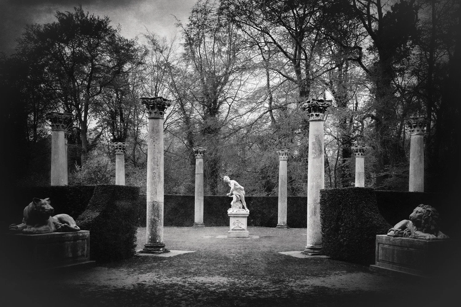 A black-and-white photograph of a classical sculpture garden featuring seven tall, classical columns, a statue in the center, and two reclining lion sculptures on rectangular bases on either side, surrounded by leafless trees and bushes.