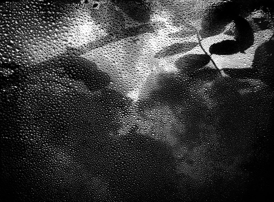 Close-up of a women's face with wet skin, seen through a rain-covered window with water droplets, reviving a mysterious and emotional mood.