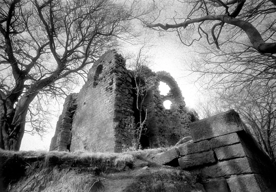 Black and white photo of the Banqueting House at Hackfall in Yorkshire,  with large trees surrounding it, some of the walls partially collapsed and holes in the structure.