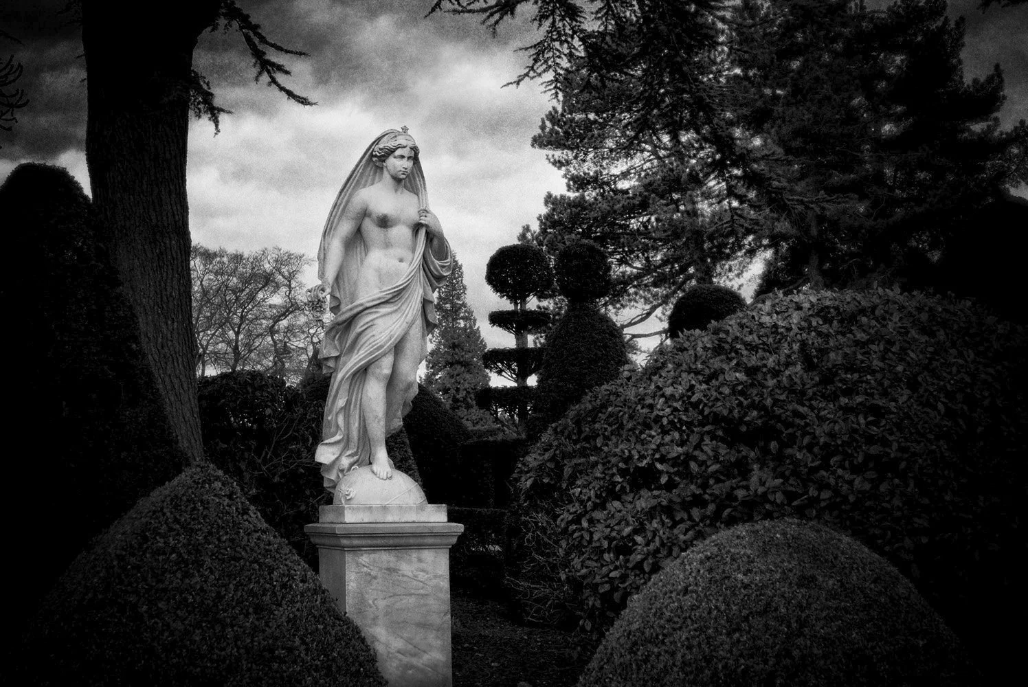 A black-and-white photograph of a classical marble sculpture of a woman, standing outdoors among trees and bushes, with a cloudy sky in the background.