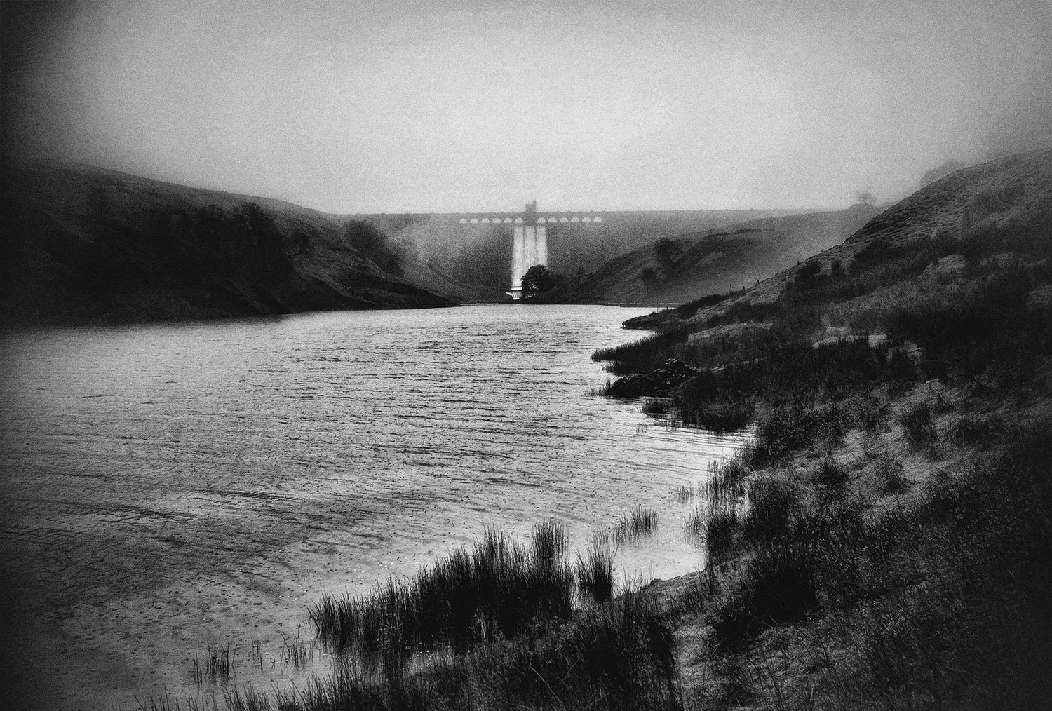 An atmospheric Black and white photo of a dam seen across a rain spattered reservoir.