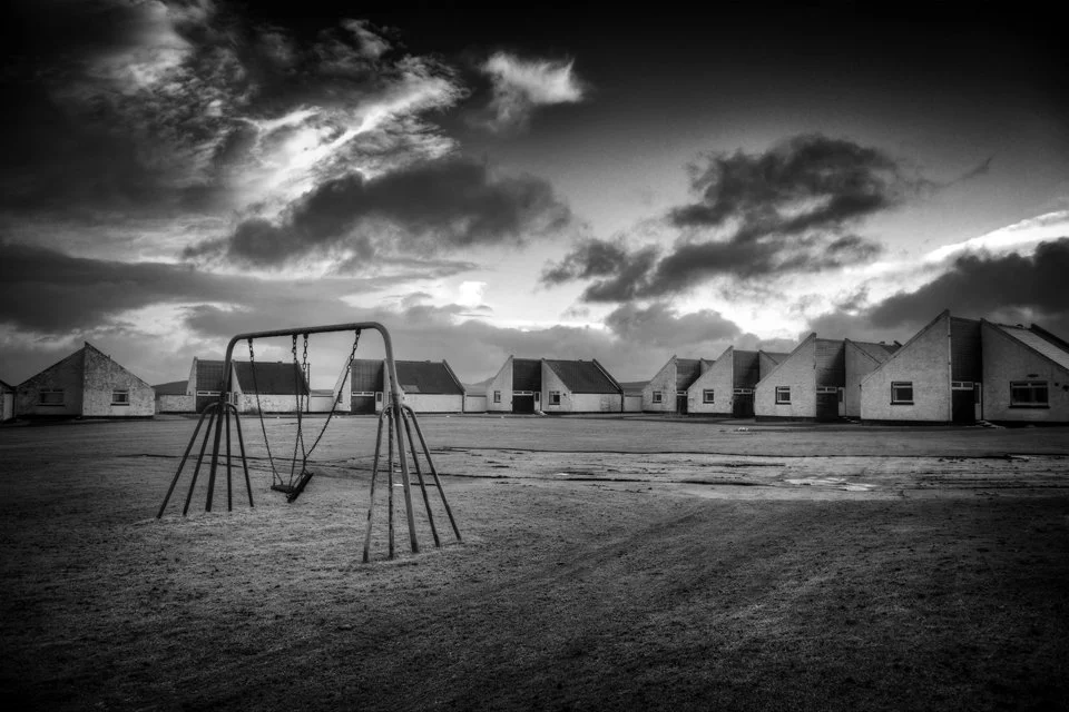 Black and white photo of a deserted playground with a swing set in front of a row of house-like structures under a dramatic cloudy sky.