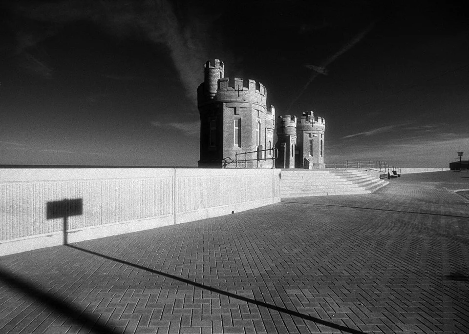 Black and white photo of a castle-like structure on a paved area with stairs, a fence, and a shadow of a sign on the ground, under a mostly clear sky with some clouds.
