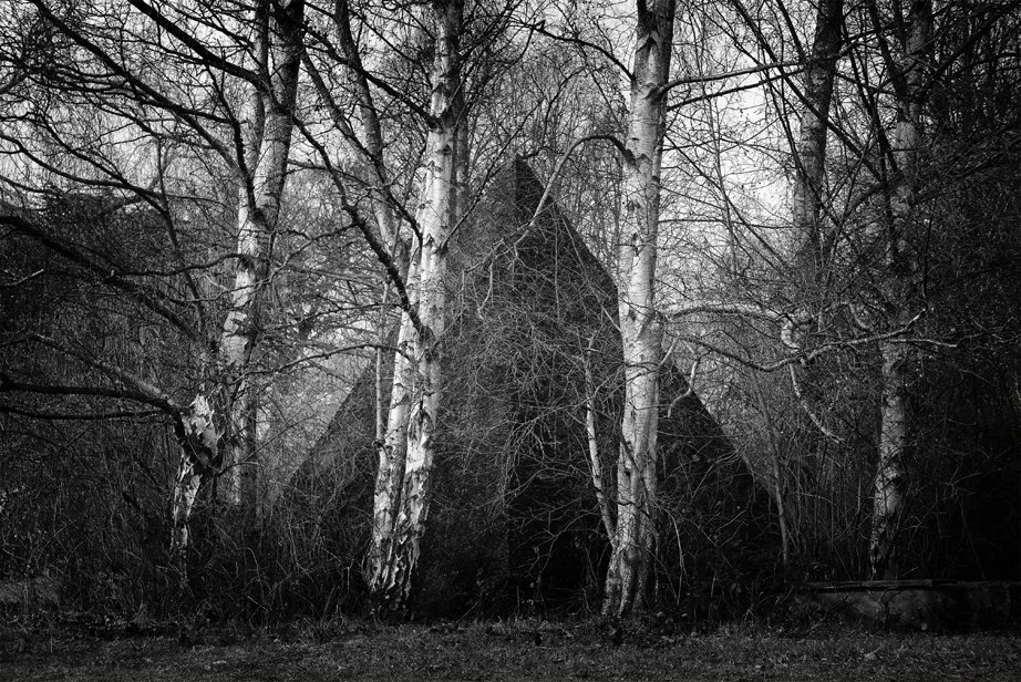 A black and white photo of a Pyramid at the Yorkshire Sculpture Park  partially obscured by leafless trees in a wooded area.