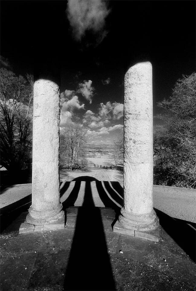 Black and white photo of two large stone columns with long shadows cast between them, overlooking a rural landscape with trees, a wooden fence, and a cloudy sky.