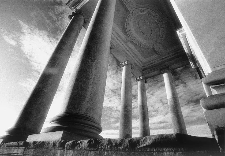 Black and white photo of classical columns on the Ionic Temple at Rievaulx Terrace, supporting a decorated ceiling, viewed from below, with a cloudy sky in the background.