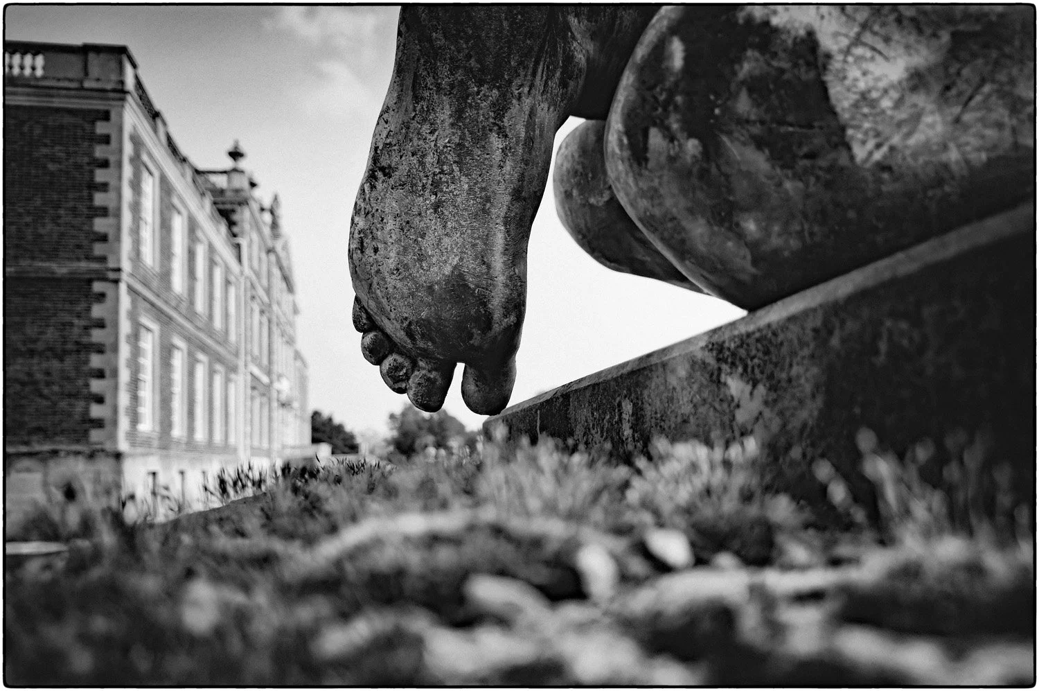 Close-up of an outdoor sculpture of a human foot hanging over a ledge with a historic brick building in the background, black and white.